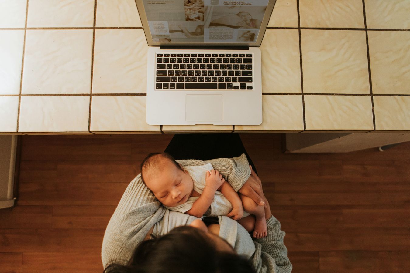 Overhead view of newborn baby in mother's arms while she sits at a table with an open laptop