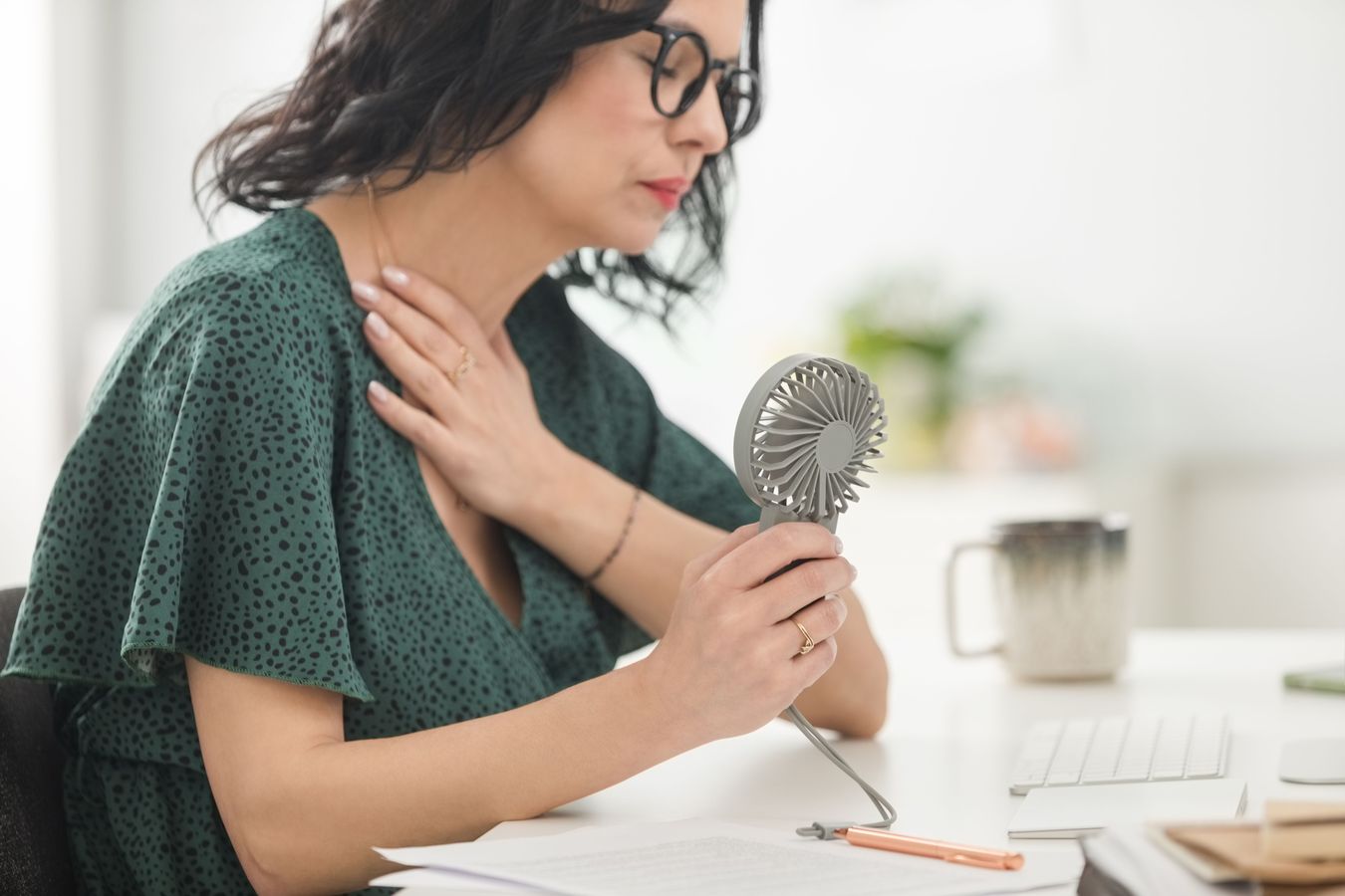 A woman holds a fan to her face while working at a desk. 